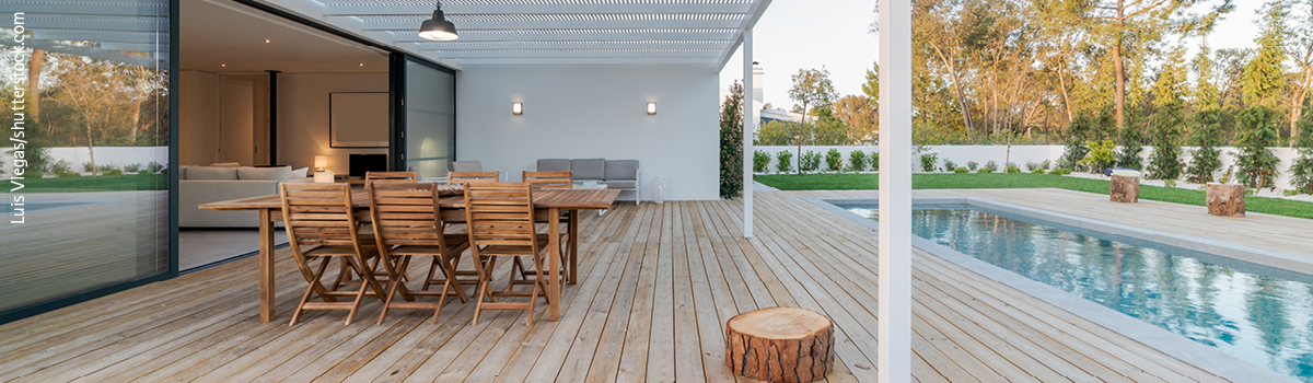 Helle holzfarbene Terrasse mit Holztisch und sechs Holzstühlen, einer Baumscheibe als Dekorationselement und einer weißen Überdachung - Pool rechts im Bild, links Zugang von hellem Wohnzimmer auf die Terrasse